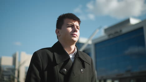 confident businessman in black coat walking outdoors under bright sunlight in urban area near modern glass buildings, looking determined against clear blue sky