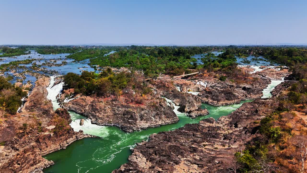 Rushing green waters of the Mekong River carve through rocky terrain and forested islets in Don Det, Laos, creating dramatic rapids and small waterfalls under a clear blue sky in this vibrant scene
