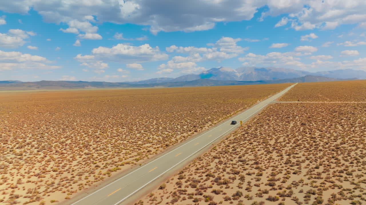 Lonely black car riding by the dry road from California to Nevada. Amazing blue skies with cotton clouds over mountains at backdrop.