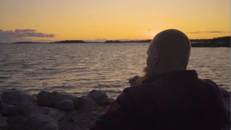 adult caucasian male with ginger beard sitting by himself on rocky beach wearing red and black shirt no hair looking over the horizon of the vibrant sunset thinking about life smoking antique pipe