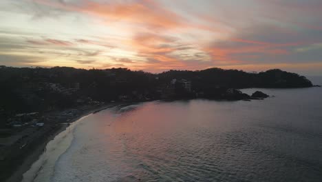 Drone-Se-Eleva-Sobre-Los-Surfistas-Atrapando-Olas-En-La-Bahía-De-Arena-Al-Atardecer,-Sayulita-México