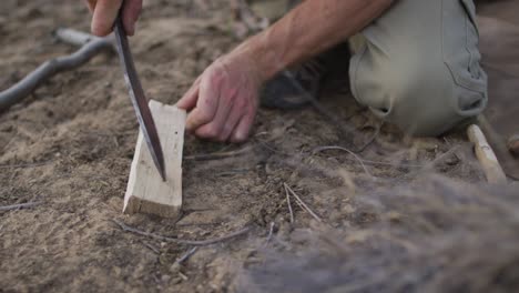 Caucasian-male-survivalist-using-machete-to-prepare-fireboard-at-camp-in-wilderness