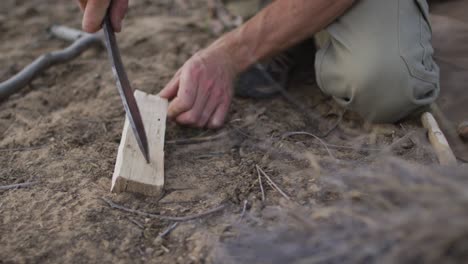 Caucasian male survivalist using machete to prepare fireboard at camp in wilderness