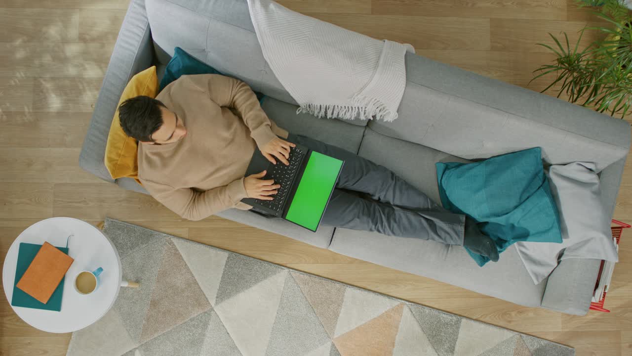 Young Man in Brown Jumper and Grey Jeans is Lying Down on a Sofa, Using a Laptop with Green Screen. Cozy Living Room with Modern Interior with Plants, Table and Wooden Floor. Top Down.