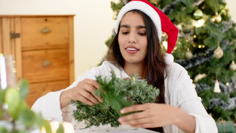 indian woman crafting christmas wreath at home, wearing santa hat, smiling warmly