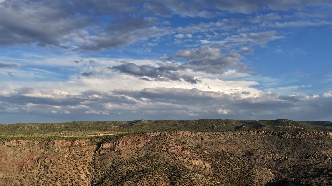 Un timelapse cinematografico con drone che rivela il bordo di un maestoso canyon desertico. Nuvole atmosferiche si evolvono dinamicamente sopra l'aspro altopiano, proiettando bellissime ombre mutevoli attraverso la vasta natura selvaggia.