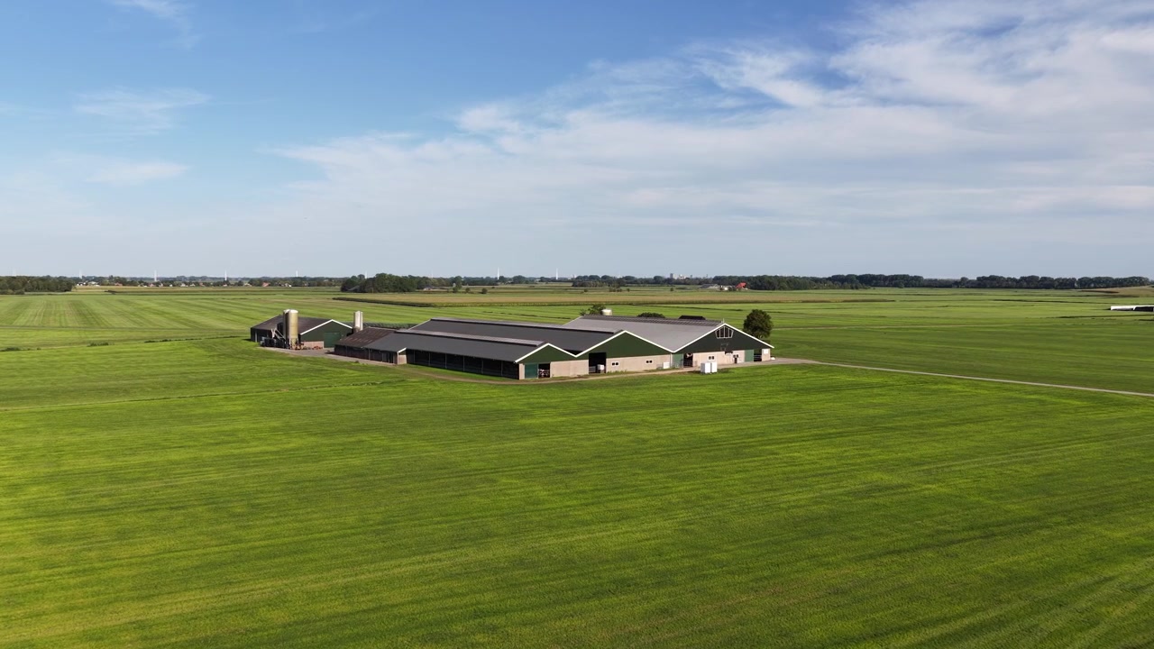 Moderne landbrugsgård med lader og siloer, der står midt i store grønne dyrkede marker under en klar blå himmel, hvilket repræsenterer bæredygtigt landbrug og fødevareproduktion.