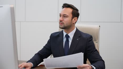executive or businessman reviews paper documents, checking computer display, nodes with positivity and smile
