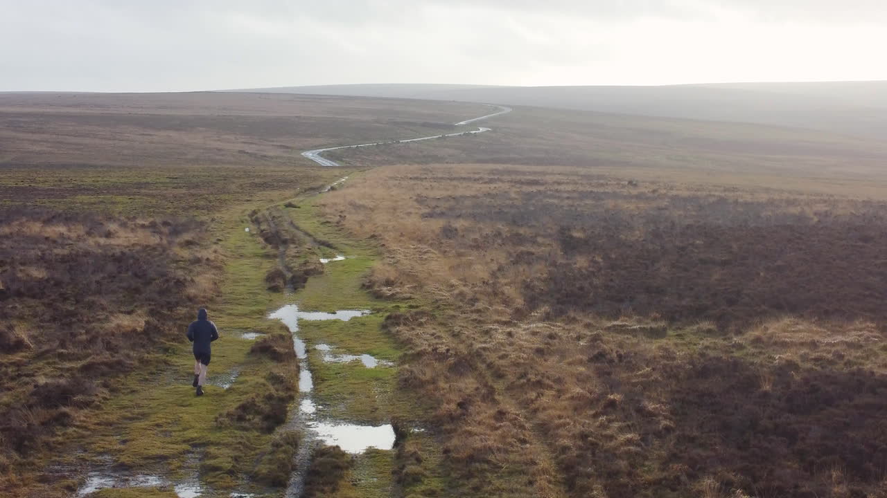 atleta corredor entrenamiento físico lado tiro aéreo drone con destello de sol en charco y páramos de campo abierto en exmoor uk 4k