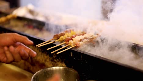 hands cooking skewered meat over smoky barbecue