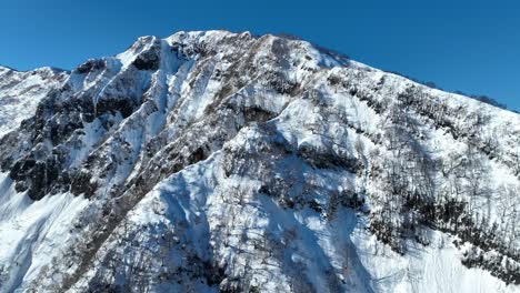 Plano-Amplio-Ascendente-Volando-Por-La-Cara-Del-Acantilado-De-Una-Montaña-Cubierta-De-Nieve,-Estableciendo-Un-Plano-Que-Revela-Enormes-Montañas