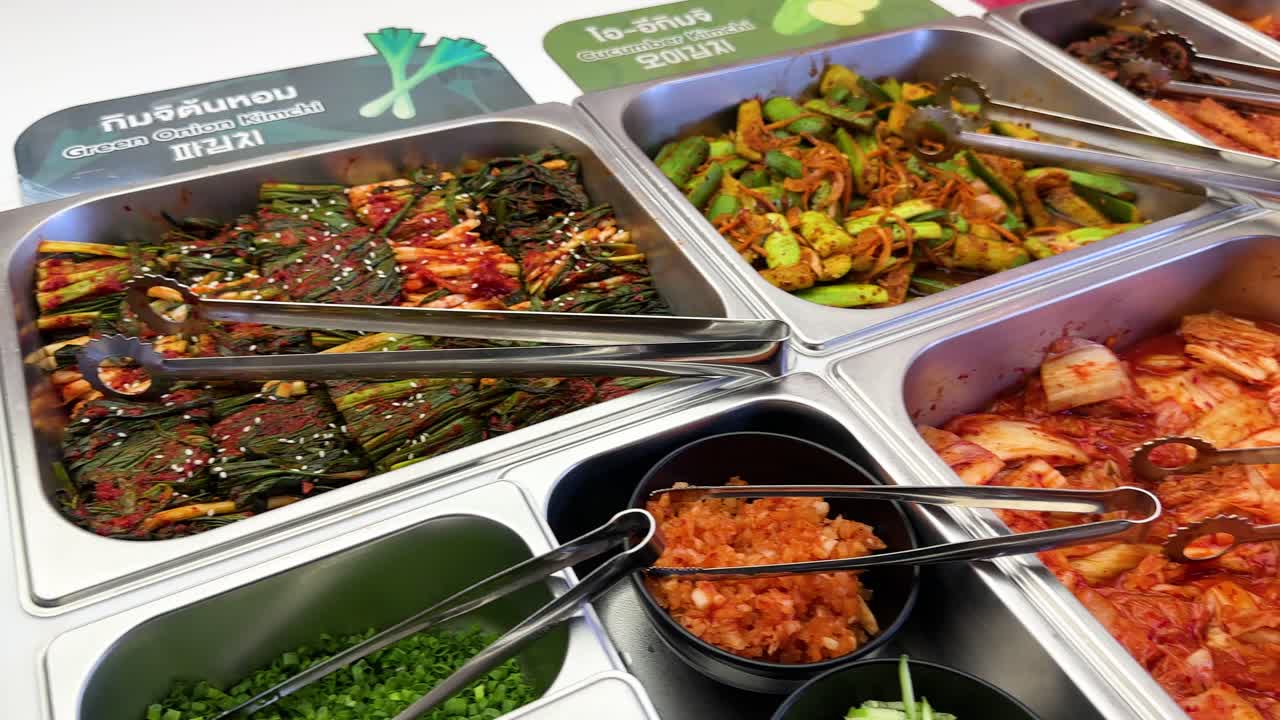 Hand chooses assorted Korean side dishes under bright lighting, buffet-style, with close-up camera movement