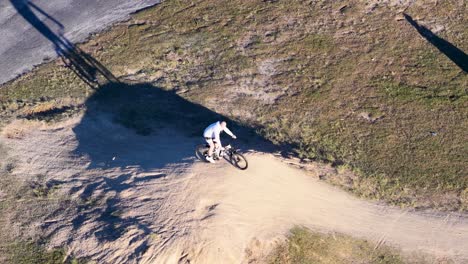 a cyclist performs stunts on a dirt track under bright sunlight, casting long shadows on the ground