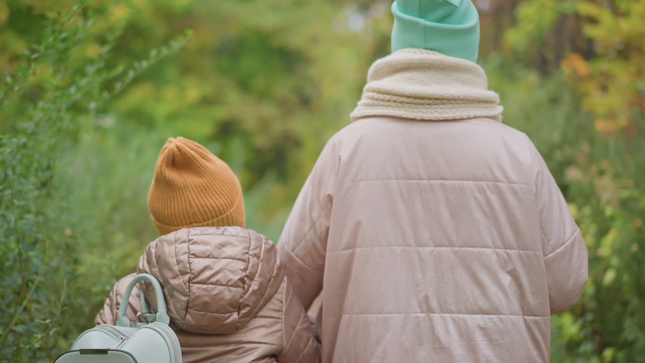 child in brown jacket and orange beanie walks beside adult in pale pink coat through lush autumn forest while holding dog leash, surrounded by soft green foliage and tranquil atmosphere