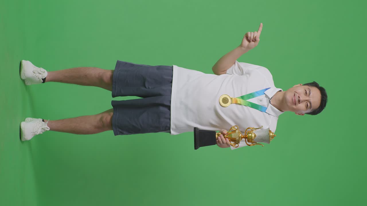 손에 금 트로피를 들고, 미소 짓고, 스튜디오의 녹색 화면 배경에 을 가리키는 금메달을 가진 아시아 남자의 몸이 가득 차 있습니다.