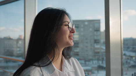 close up of cheerful woman with long hair and glasses holding tablet while smiling and looking outdoors through large glass window, sunlight highlighting her face with urban buildings in background