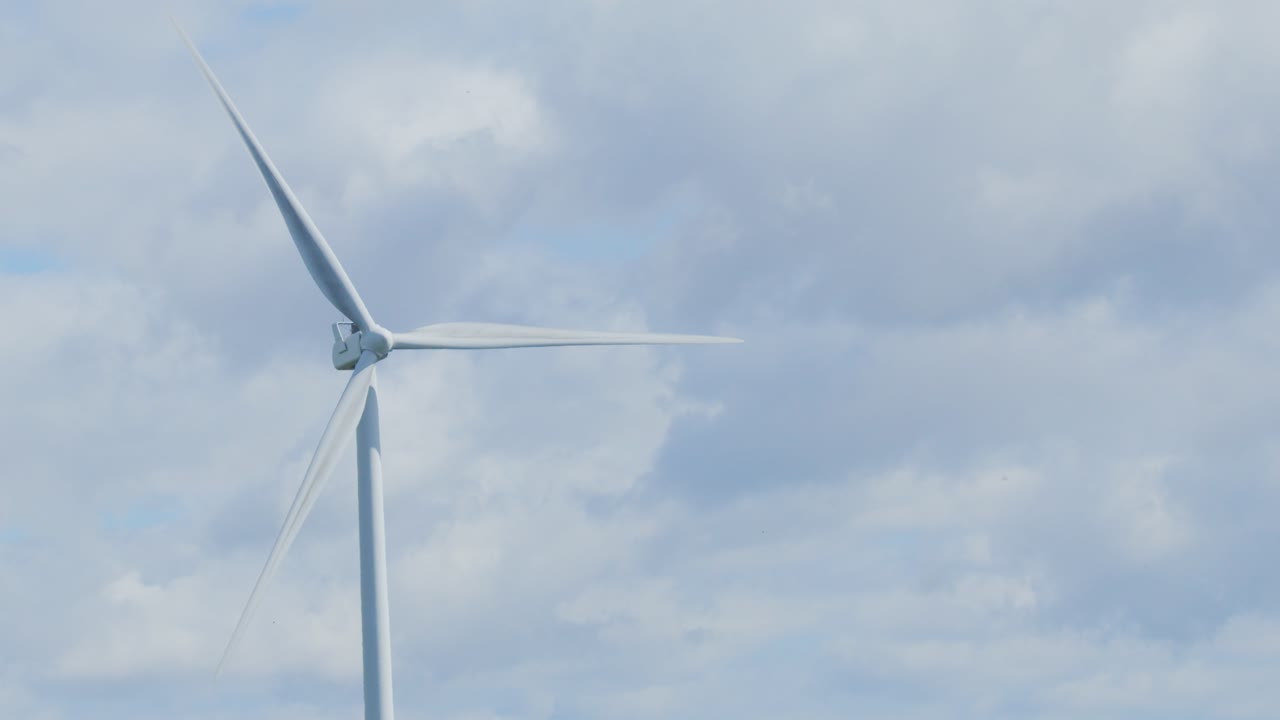 A single modern wind turbine spins steadily in an open landscape under a bright, overcast sky. Static camera, wide shot, natural daylight