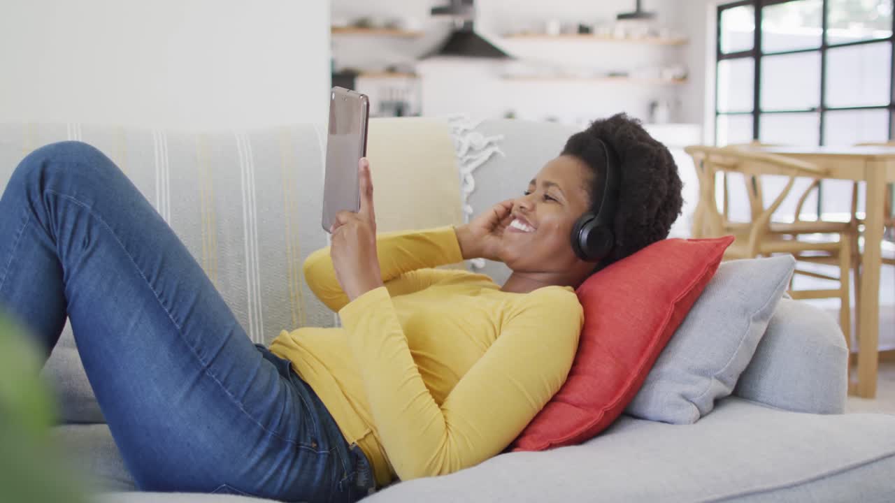 Happy african american woman wearing headphones laying on sofa using tablet