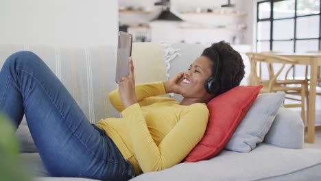Happy-african-american-woman-wearing-headphones-laying-on-sofa-using-tablet