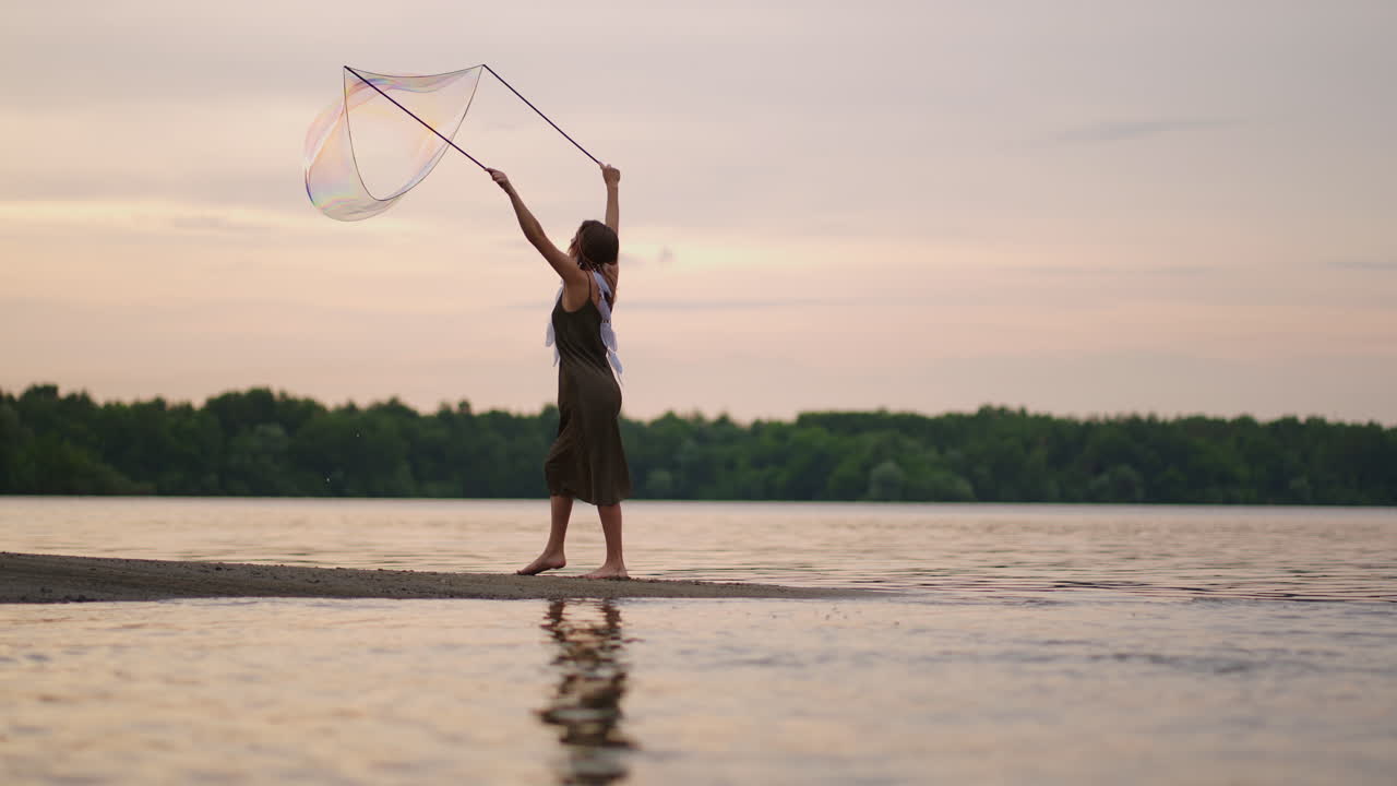 젊은 여성 예술가 는 일몰 에 호수 연안 에 거대한 비누 거품 을 불어넣는 비누 거품 쇼 를 보여준다. 느린 움직임 으로 비누 거품 의 아름다운 쇼 를 보여준다