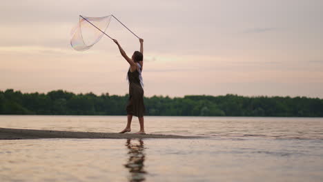 a young female artist shows a soap bubble show blowing up huge soap bubbles on the shore of a lake at sunset. show a beautiful show of soap bubbles in slow motion