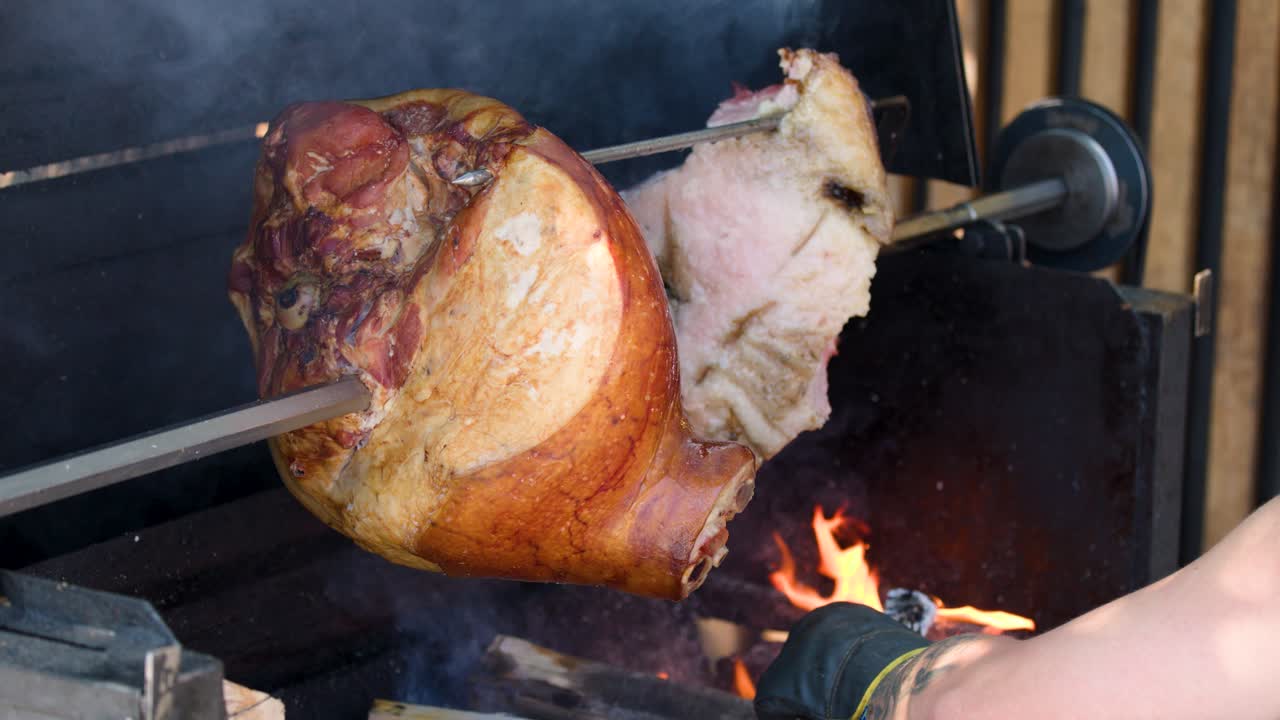 Gloved hand rotates large Prague ham on spit above open flames, outdoor market setting