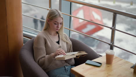 thoughtful lady flips to new page while reading book in cozy cafe with city view seen through glass panel, coffee cup and phone placed on table in front of her