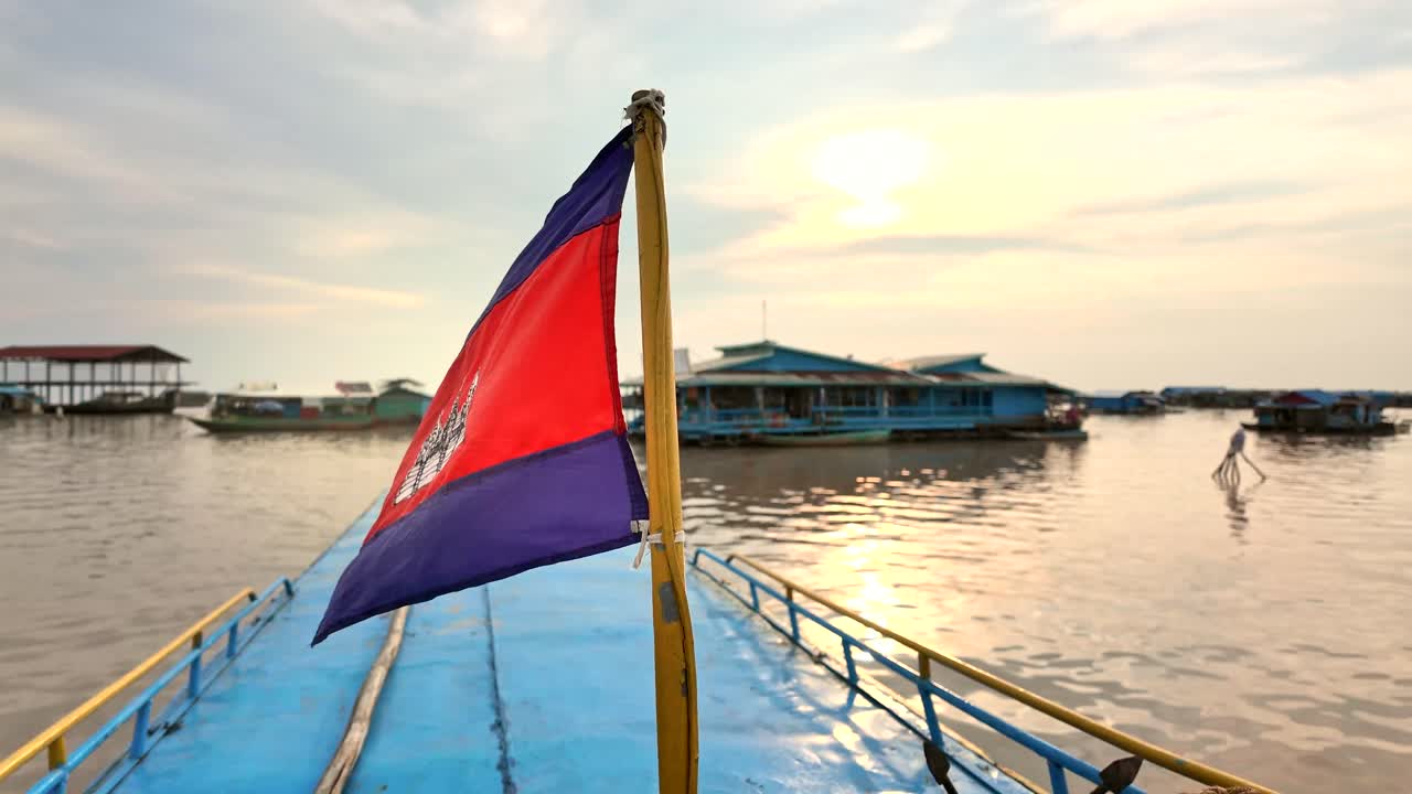 Cambodian flag waves atop a blue boat on Tonle Sap Lake, stirred by the evening breeze. Golden sunset light reflects on the rippling water, while a floating village rests in the background.