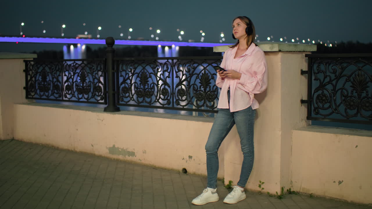 Young lady in pink top and headphones resting on the fence with a peaceful vibe, illuminated bridge lights in the background with cars moving on the bridge, and a relaxed evening atmosphere
