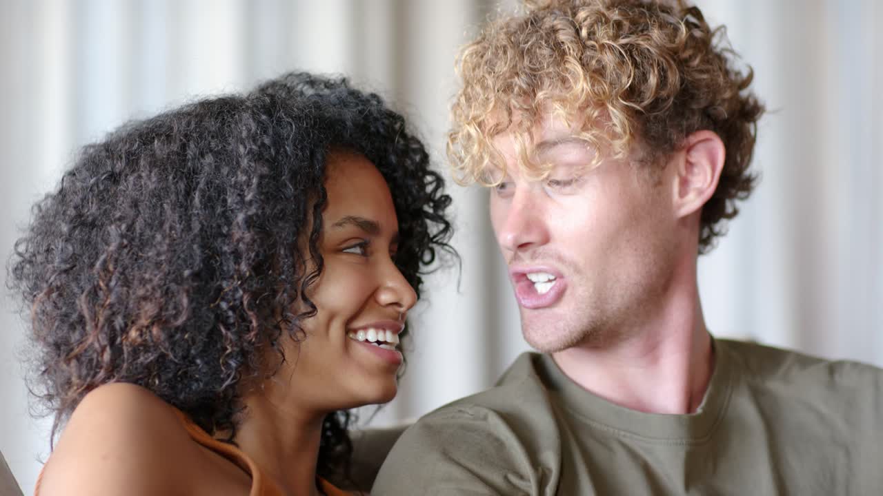 Diverse couple sharing warmth, woman touching man's arm, man making faces, laughing on sofa