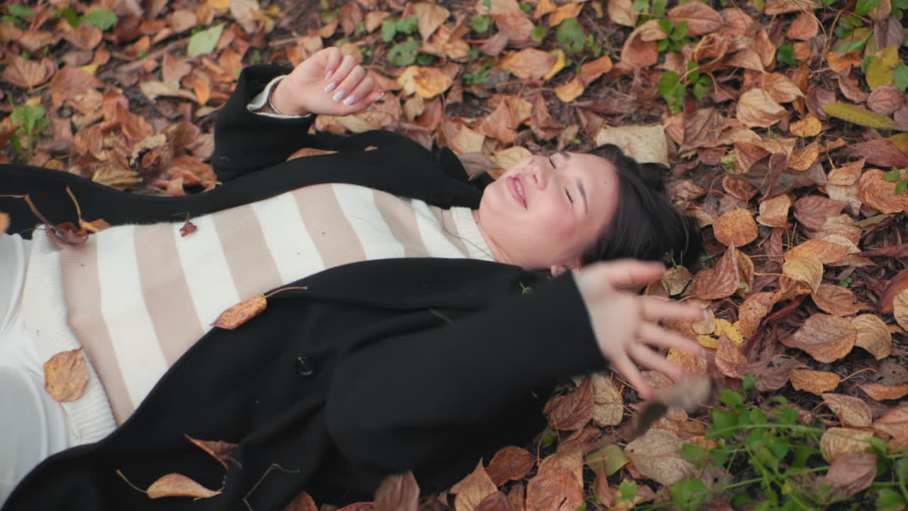 Free spirited girl lies face up on autumn forest floor, smiling while blocking falling dry leaves with hands, cozy coat and striped sweater visible, playful mood under soft light