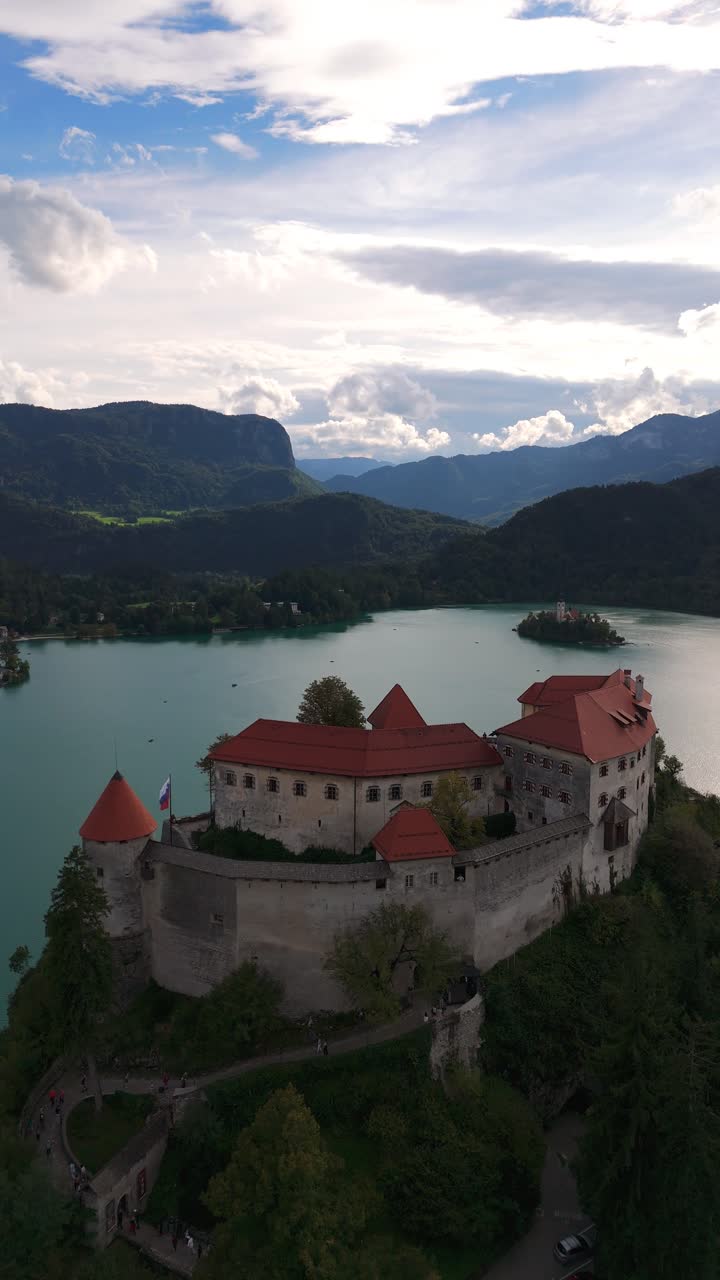 Drammatico primo piano verticale aereo del castello di Bled arroccato sopra il lago circondato da montagne verdi, Slovenia