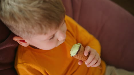 close up top down view of young boy in orange shirt holding and enjoying ice cream cone while seated indoors on sofa, capturing quiet childhood