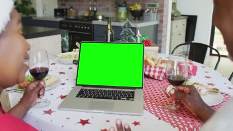una pareja afroamericana sonriente con sombreros de santa haciendo una llamada de video de navidad con computadora portátil, con pantalla verde