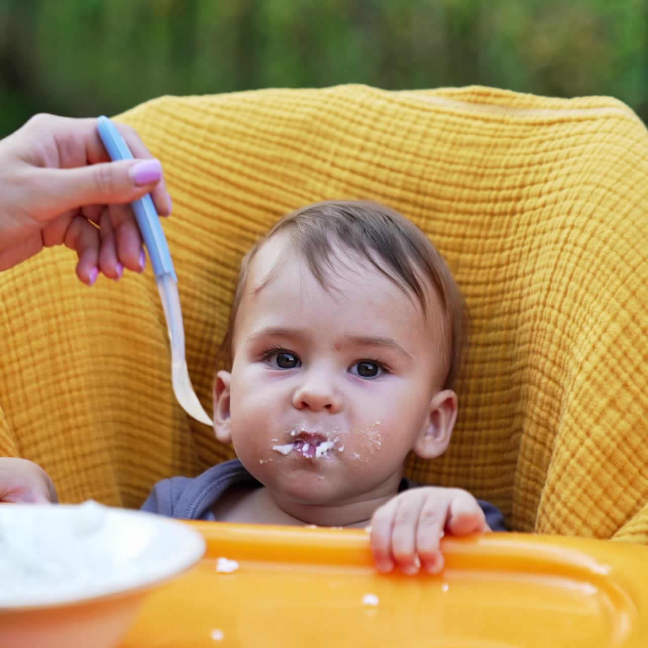 Loving mom giving food from spoon to a baby in yellow chair. Baby nutrition outdoors in the garden. Blurred nature backdrop