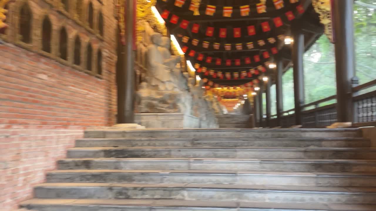 Point de vue de marche le long d'un couloir de temple à Ninh Binh, montrant des statues de pierre alignées le long des marches, des drapeaux suspendus, des murs sculptés et un éclairage chaleureux à l'intérieur d'un complexe bouddhiste sacré.