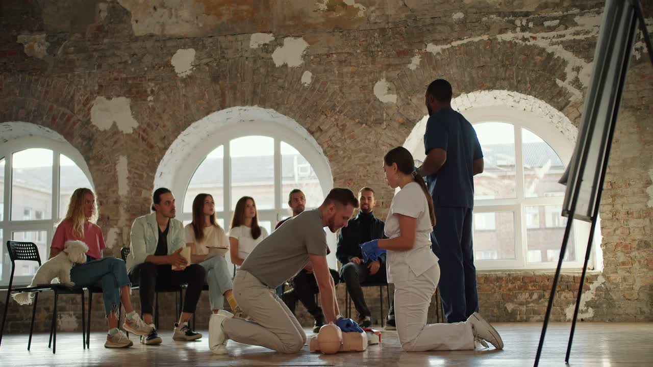 회색 티셔츠를 입은 수염이 있는 갈색 남자가 인공 호흡을 시키고, 색 유니폼을 입은 간호사 소녀가 흑인과 함께 파란 유니폼에 입은 의사와 함께 벽돌 벽과 큰 창문의 배경에서 대중 앞에서 응급 처치 규칙을 설명합니다.