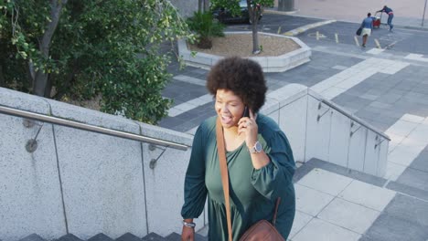 happy plus size biracial woman walking on stairs and talking on smartphone in city