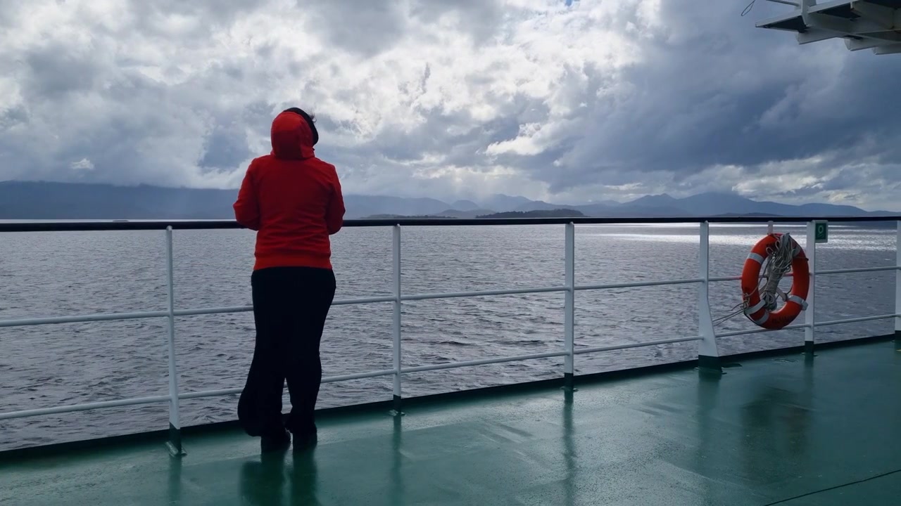 Una donna si appoggia alla ringhiera del traghetto durante un viaggio attraverso i fiordi della Patagonia cilena, vivendo un viaggio veloce, freddo e ventoso circondata da paesaggi selvaggi e drammatici e natura incontaminata.