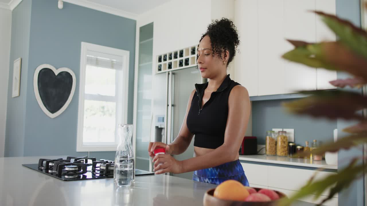 Donna afroamericana che prende una caraffa in cucina, versando, e prendendo pillole dalla bottiglia per la salute.