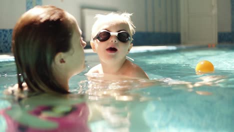 Young-woman-holding-his-son,-teaching-to-swim.-Supporting-above-the-water.-Indoors-swimming-pool.-Little-boy-in-swimming-glasses