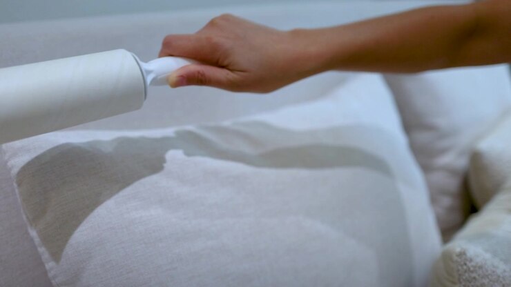 Primo piano di una mano che pulisce un cuscino in tessuto con un rullo levapelucchi in un ambiente luminoso della camera da letto.