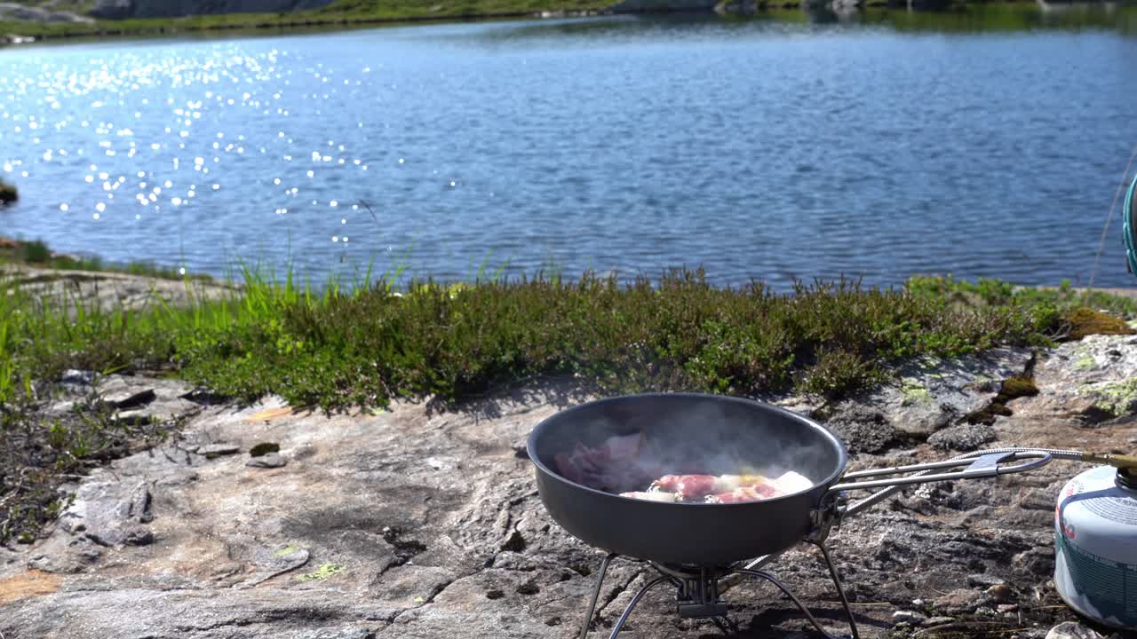 persona cocinando huevos y tocino en un pequeño quemador mientras acampa en un pintoresco lago de montaña