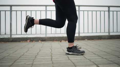 leg view of athlete in black sneakers and leggings jogging near iron railing of bridge overlooking partial foggy cityscape during winter