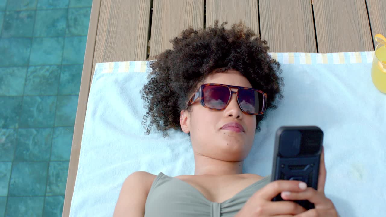 African American woman lying on towel on wooden pool deck, holding flip phone, checking messages