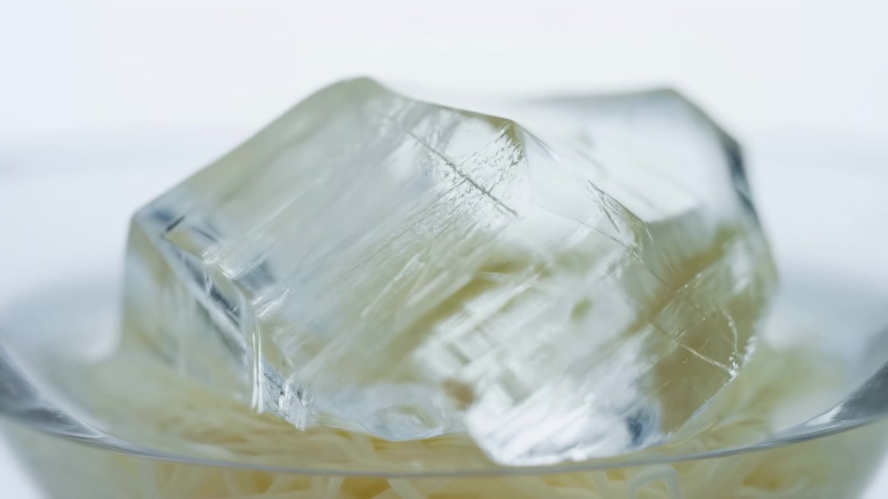 透明なガラスボウルの縁に置かれた大きな半透明の氷の塊のクローズアップタイムラプス。最初はほぼ完全に固形だったものが、ほぼ完全に溶けるまで進行する様子。