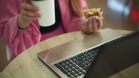 hand view of a woman working on her laptop with a partially eaten burger in her left hand, pausing to take her drink from the table, placing it back, and continuing her work, background slightly blur