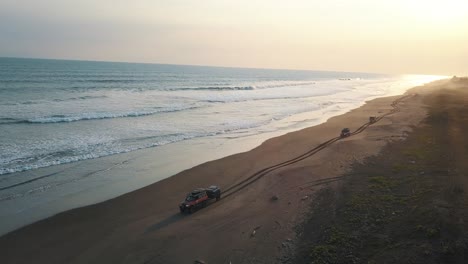 drone aerial of 4x4 cars driving on the shore of black sand beach during sunset