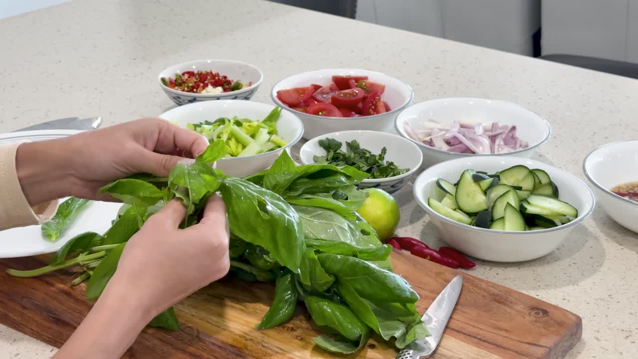 A person hand-picks fresh basil leaves on a kitchen counter, surrounded by bowls of chopped vegetables, under bright, even lighting with a steady camera angle
