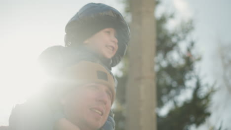 close-up of a father carrying his joyful son on his shoulders, as the boy playfully moves up and down, the father walks focused, while the warm sunlight casts a bright glow over them